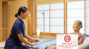 ベッドに横たわる高齢女性に対して、紺色のユニフォームを着た看護師が点滴の管理をしている様子。背景は明るい和室で、点滴スタンドが見える。