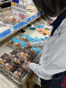 白い長袖を着た女性がスーパーのベーカリーコーナーで、青いパッケージの食パンを手に取っている様子。奥には260円の価格表示が見える。