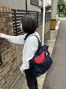 白いジャケットを着た女性が、レンガ造りの住宅の門扉に手をかけている後ろ姿。紺と赤のバッグを背負っている。