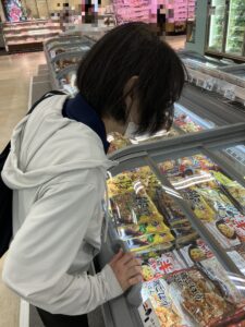 白いパーカーを着た女性が、スーパーの冷凍食品売場のケースに並ぶ商品を身を乗り出して確認している様子