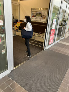 マスクを着用した女性がクリニックの入口で黄色い買い物かごを持って立っている。背後には情報ボードと待合スペースが見える。
